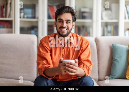Smiling Middle Eastern male holding coffee cup sitting on sofa at home Stock Photo