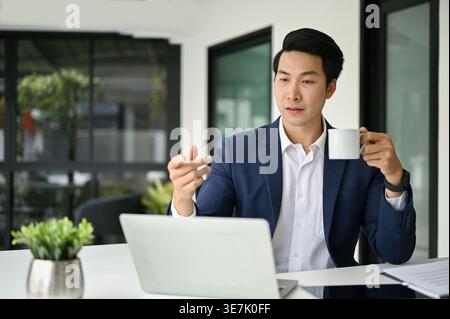 Asian businessman having online meeting on laptop holding coffee cup sitting at table in office Stock Photo