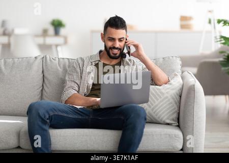 Happy young Arab man using laptop for video conference, calling on smartphone, working online from home. Millennial Eastern guy communicating to colle Stock Photo