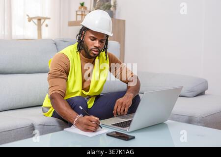 Black male engineer with twists wearing hard hat and safety vest taking notes while sitting on sofa near coffee table with laptop and smLifestylephone at... Stock Photo