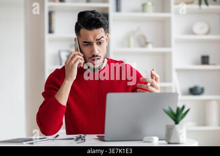 Upset Arab man talking on smartphone looking at laptop screen sitting at table at home Stock Photo