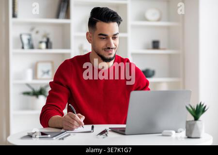 Arab man writing down notes looking at laptop screen sitting at desk at home Stock Photo