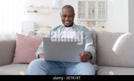 Smiling Black man using laptop while sitting on sofa at home Stock Photo