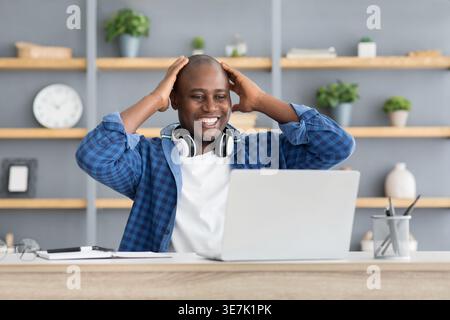 Excited Black businessman wearing headphones around neck looking at laptop screen and touching head while sitting at desk in office Stock Photo