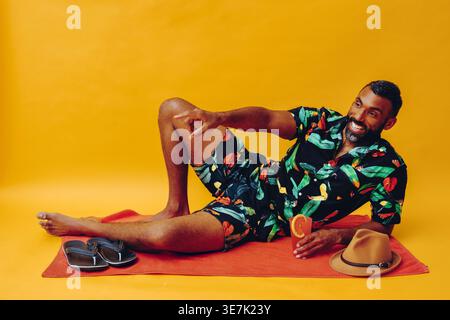 Mixed-race bearded man leaning on elbow holding orange juice pointing and looking away while lying sideways on towel with flip flops and fedora hat... Stock Photo