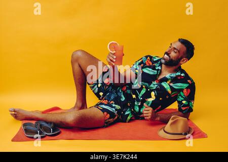 Mixed-race bearded man leaning on elbow holding and looking at orange juice while lying sideways on towel with flip flops and fedora hat against ye... Stock Photo
