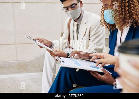 Indian businessman with eyeglasses and face mask discussing reports with Black female colleague sitting outdoors during daytime Stock Photo