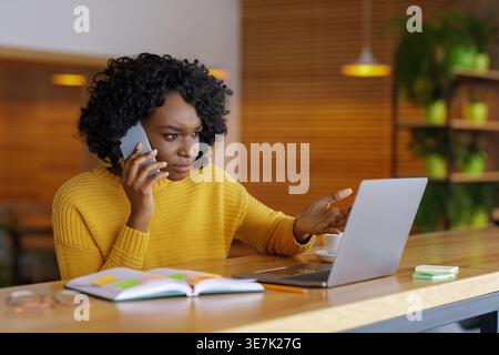 Serious Black woman with curly hair gesturing hand having phone call looking at laptop sitting at table with notebook in cafe Stock Photo