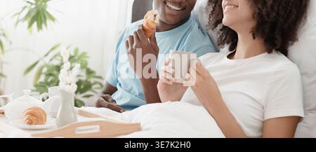Smiling Black couple using smartphone having breakfast while sitting on bed in bedroom at home Stock Photo
