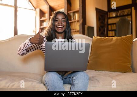 Black teenage female with laptop showing thumbs up sitting on couch in living room at home Stock Photo