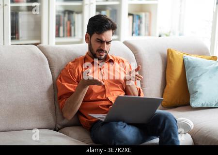 Irritated Middle Eastern man gesturing hands having video call using laptop sitting on couch at home Stock Photo