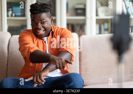 Smiling Black man with braids with crossed arms pointing finger forward while recording video sitting on sofa in living room at home Stock Photo