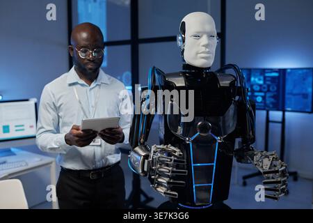 Black male computer engineer with eyeglasses using tablet while standing near robot in laboratory Stock Photo
