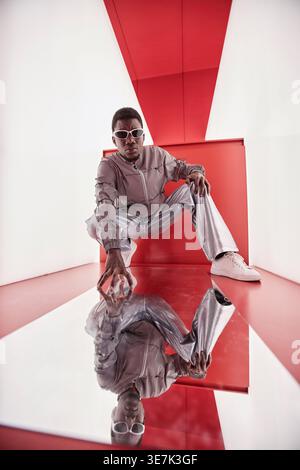 Black man wearing sunglasses and reflective outfit while crouching against red and white background in studio Stock Photo