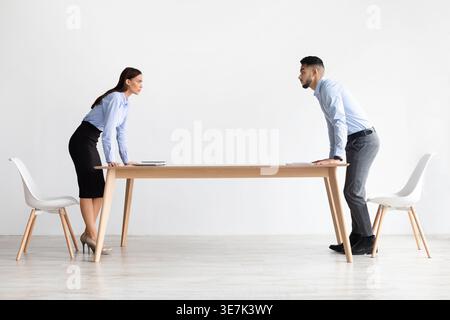 Conflict At Work. Two Angry Young Businessman And Businesswoman Staring At Each Other Arguing Having Confrontation Standing At Table In Modern Office, Stock Photo