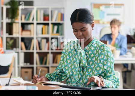Black female student looking at tablet screen and writing on notebook while sitting at table near caucasian female teacher in classroom Stock Photo