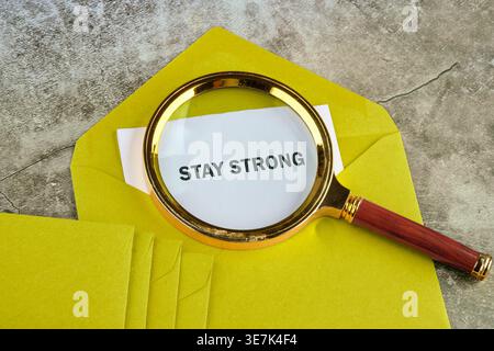 Magnifying glass rests on an open yellow envelope, enlarging the words Stay Strong on white paper. Multiple yellow envelopes stack nearby on a grey su Stock Photo