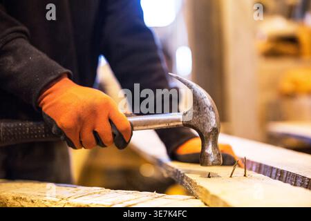 Hands of mixed-race male teenager carpenter wearing gloves using hammer on wood while standing in workshop Stock Photo