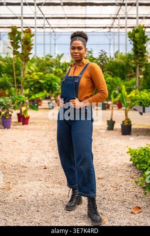 Hispanic female gardener in denim overalls standing in greenhouse during daytime Stock Photo