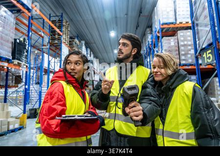 Diverse group of warehouse workers wearing safety vests and warm clothing using barcode scanner and clipboard while standing and inspecting product... Stock Photo