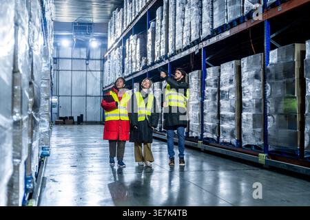 Diverse group of logistics team in safety vests discussing inventory management while walking with barcode scanner and clipboard in cold storage wa... Stock Photo