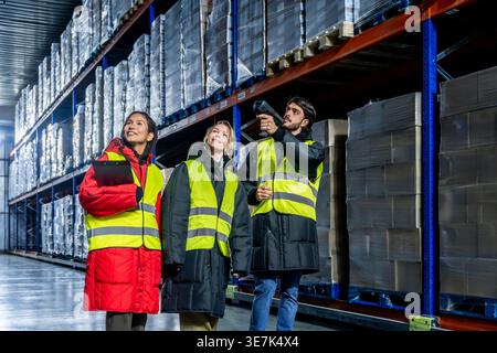 Diverse group of logistics team in safety vests discussing inventory management while walking with barcode scanner and clipboard in cold storage wa... Stock Photo
