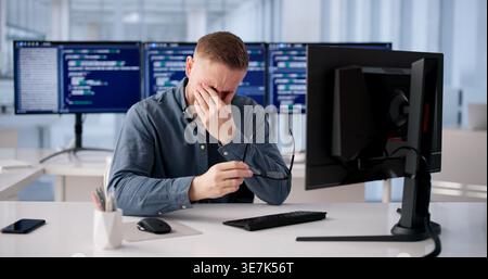 Tired Coder Rubs Irritated Eye During Extended Hours At Workstation Stock Photo