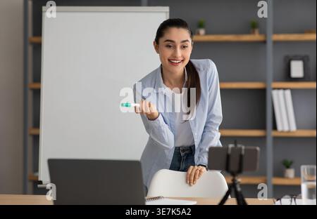 Arab female tutor holding marker explaining lesson at whiteboard teaching online using smartphone while standing near table with laptop at home Stock Photo