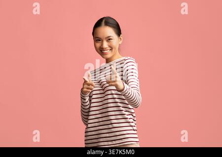 Smiling Asian female pointing fingers forward standing against pink background in studio Stock Photo