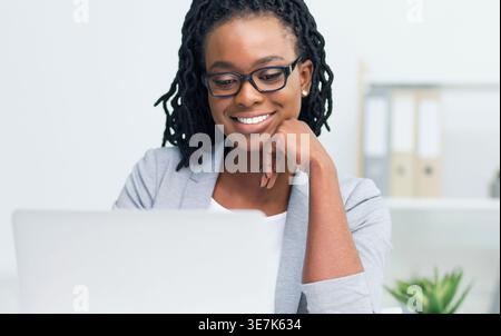 Smiling Black business woman with twists wearing gray coat and eyeglasses looking at laptop while sitting at table in office Stock Photo