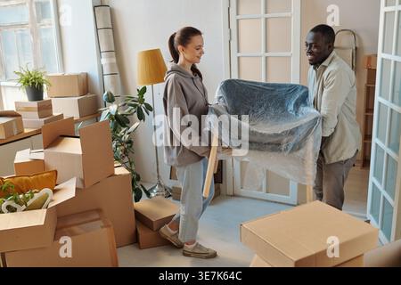 Black man standing and carrying new armchair with caucasian girlfriend during relocation in their new home Stock Photo