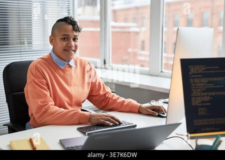 Smiling mixed-race female programmer sitting at table with computer and laptop in office Stock Photo