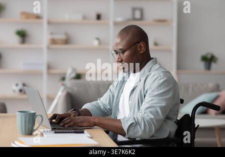 Cheerful disabled Black bald businessman in wheelchair wearing eyeglasses working on laptop at table with smartphone and documents in living room a... Stock Photo