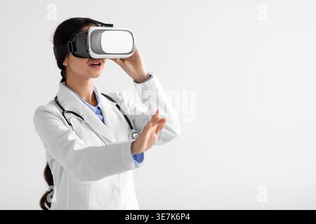 Indian female doctor with stethoscope on neck wearing VR glasses experiencing virtual reality while standing against white background in studio Stock Photo
