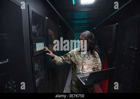 Black female soldier wearing eyeglasses holding laptop checking cabinet monitor while standing in server room Stock Photo