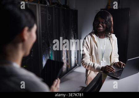 Black female IT engineer wearing eyeglasses talking to caucasian female colleague holding tablet standing at table with laptop in server room Stock Photo