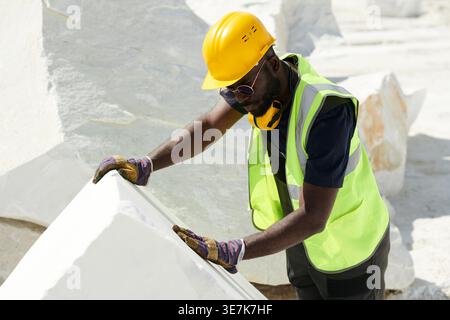 Black male worker wearing safety vest and hard hat inspecting cube of rock while standing in construction site on sunny day Stock Photo
