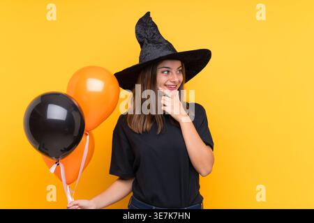 Hispanic woman in halloween costume looking sideways touching chin holding black and orange balloons standing against yellow background in studio Stock Photo