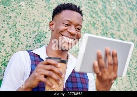Black man holding takeaway coffee and using tablet while standing in front of concrete wall outdoors Stock Photo