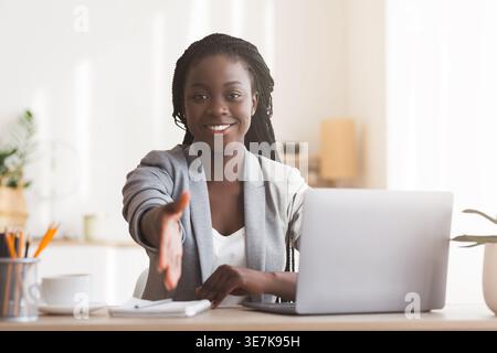 Black woman sitting at desk with open laptop hand reaching out for handshake in the office during daytime Stock Photo