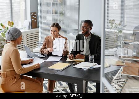 Mixed race female candidate wearing headwrap and eyeglasses holding resume sitting at table with male and female HR managers discussing employment ... Stock Photo