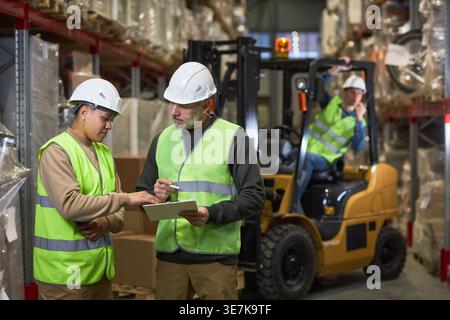 Mixed-race female and caucasian male workers holding clipboard and talking while standing in warehouse Stock Photo