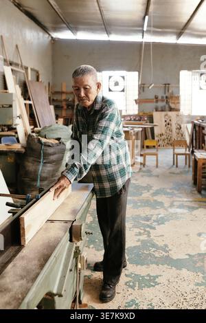 Senior Asian male carpenter holding wooden board standing at workbench while working in workshop Stock Photo