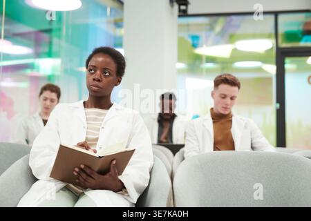 Black female medical student in lab coat taking notes while listening to lecture sitting in audience during medical seminar in conference room Stock Photo