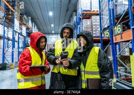 Diverse group of warehouse workers wearing safety vests and warm clothing using barcode scanner and clipboard while standing and inspecting product... Stock Photo