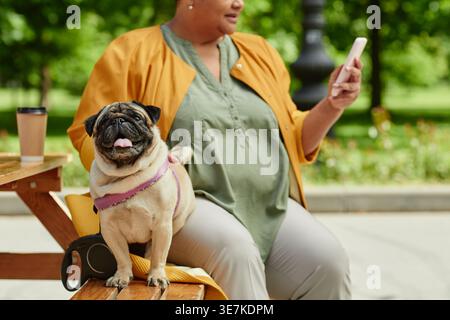 Cropped shot of Black middle-aged woman with her dog sitting on wooden bench using smLifestylephone in outdoor cafe Stock Photo