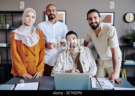 Middle Eastern businessman with laptop sitting at table with colleagues standing around him in office Stock Photo