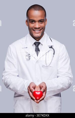 Smiling black doctor with stethoscope around neck holding heart shape toy while standing against grey background in studio Stock Photo