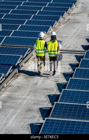 Hispanic and caucasian female engineers wearing hard hats and safety vests discussing work while walking on commercial rooftop with solar panels on... Stock Photo