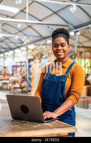 Hispanic female employee managing orders using laptop while standing at wooden table in garden center Stock Photo